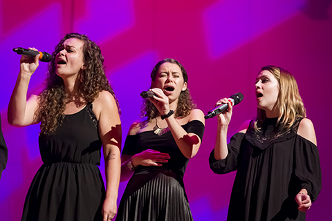 Three vocalists in black dresses hold up microphones as they perform on stage